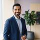 A professional portrait of a South Asian man smiling, set against a modern, soft-toned studio background.