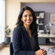 A professional portrait of a smiling South Asian woman in a modern, well-lit office setting.