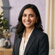 A friendly South Asian woman smiling in a professional headshot, set against a soft lavender and cream studio background.