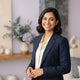 A professional portrait of a smiling South Asian individual in a modern, light-filled workspace with lavender and cream tones